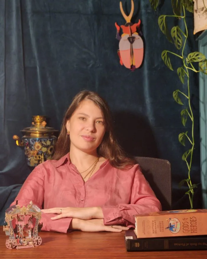 Masha Kartashev seated at her desk in her office, hands folded, with a plant and pop-up books beside her
